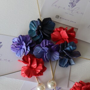 May include: A selection of fabric flower lapel pins in various colours, including red, blue, and purple. Each pin has a gold-coloured stem and a pearl detail. The pins are presented with business cards bearing the text "lavann.dana".