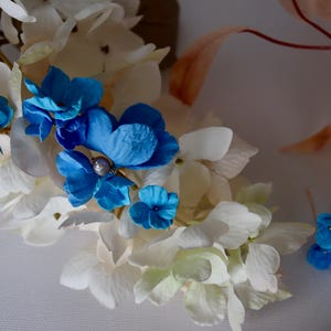 Puede incluir: Primer plano de un arreglo floral con flores artificiales blancas y azules. El arreglo presenta grandes flores de hortensia blancas y flores azules vibrantes. Una pequeña perla está en el centro de una flor azul.