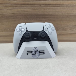 Puede incluir: Un mando blanco y negro de PlayStation 5 sobre un soporte blanco. El soporte muestra "PS5" con un logotipo de corona. El mando tiene joysticks y botones negros. El fondo es de madera marrón claro.