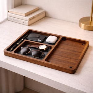 May include: A wooden desk tidy with several compartments. It contains a pen, wallet, sunglasses, watch, and wireless earbuds. The organiser is made of dark wood and is rectangular.