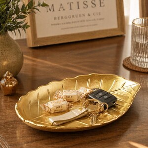 Könnte beinhalten: Eine goldfarbene, blattförmige Schale mit Schlüsseln, einer Fernbedienung und zwei verpackten Süßigkeiten. Die Schale steht auf einer Holzoberfläche, im Hintergrund ein gerahmter Druck. Eine kleine dekorative Vase und ein Glas sind ebenfalls zu sehen.