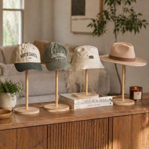 May include: Four hats displayed on wooden stands. The hats include a baseball cap with "North Dakota 1889", a green cap with "California West Coast", a white bucket hat with a bear, and a tan fedora. A candle and books are also visible.