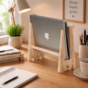 May include: A wooden laptop stand with a gray laptop. The stand is made of light-colored wood and has a simple, modern design. The laptop is positioned upright, displaying the Apple logo. The stand is on a wooden desk with a notebook, pen, and coffee cup.