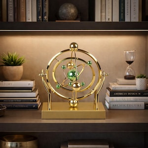 May include: A gold-colored, decorative kinetic sculpture with a spherical green center and smaller green and gold spheres. The sculpture is mounted on a rectangular base and features concentric rings, resembling a model of the solar system. It is displayed on a shelf with books and a small plant.