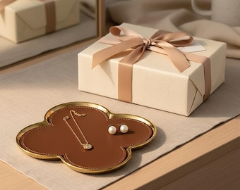 May include: A brown leather and gold-rimmed clover-shaped tray holds a gold necklace and pearl earrings. A cream-colored gift box with a satin ribbon sits nearby. The scene is set on a neutral-toned surface.
