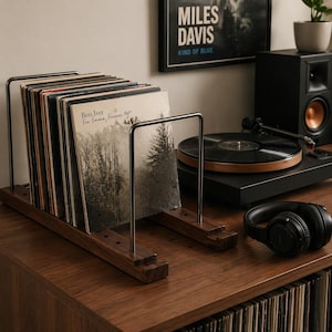 May include: A wooden record holder with metal dividers holds several vinyl records. The album covers are visible, including one by Bon Iver. A turntable, speaker, and headphones are also on the wooden surface, creating a music-themed display.