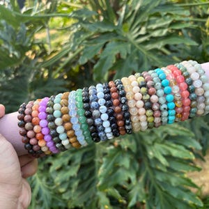 May include: A selection of beaded bracelets in diverse colours, such as brown, purple, green, and blue, are presented on a pink cylindrical holder. The bracelets are composed of small, round beads and are held by a hand against a backdrop of green foliage.