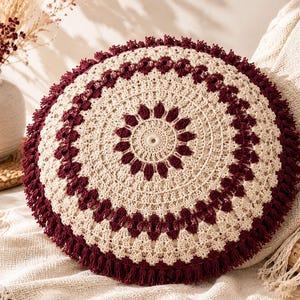 May include: A round crochet pillow featuring a circular mandala pattern in cream and deep burgundy yarn. The design includes intricate stitch work with a central floral motif and a thick, textured fringe border in burgundy surrounding the edge.