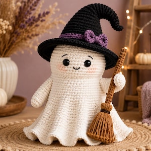 May include: A white crocheted ghost doll wearing a black witch hat with a purple bow. The ghost has black bead eyes, a small stitched smile, and pink blush on its cheeks. It holds a brown crocheted broomstick in one of its small arms.