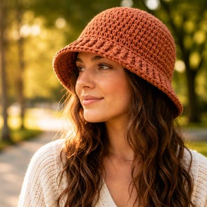 May include: A rust-colored crochet bucket hat featuring a textured stitch pattern and a slightly flared brim. The hat is constructed from yarn and displays a consistent, dense weave throughout the crown and the structured, downward-sloping brim.