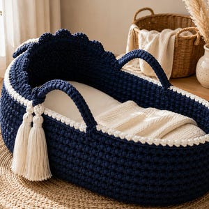 May include: A deep navy blue crocheted baby bassinet featuring a scalloped top edge, two sturdy matching handles, and two long cream-colored decorative tassels hanging from one side. The interior is lined with a soft, plain white fabric.