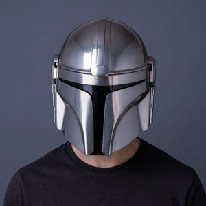 Op de afbeelding: Een zilveren Mandalorian helm met een zwart vizier. De helm heeft een glad, reflecterend oppervlak en een kenmerkend T-vormig vizier. De helm wordt gedragen door een persoon die een zwart t-shirt draagt.