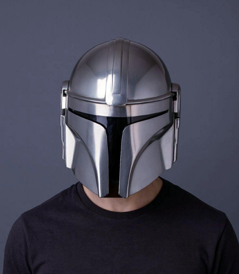 Op de afbeelding: Een zilveren Mandalorian helm met een zwart vizier. De helm heeft een glad, reflecterend oppervlak en een kenmerkend T-vormig vizier. De helm wordt gedragen door een persoon die een zwart t-shirt draagt.