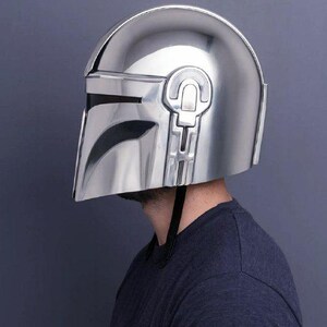 Op de afbeelding: Een glanzende, zilveren Mandalorian helm, met een T-vormig vizier en opvallende oorstukken. De helm wordt gedragen door een persoon die een donkerblauw shirt draagt. Het oppervlak van de helm is glad en reflecterend.