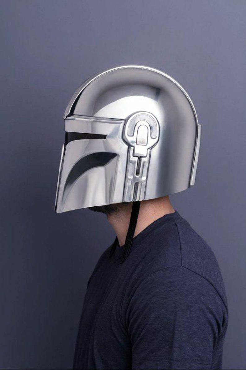 Op de afbeelding: Een glanzende, zilveren Mandalorian helm, met een T-vormig vizier en opvallende oorstukken. De helm wordt gedragen door een persoon die een donkerblauw shirt draagt. Het oppervlak van de helm is glad en reflecterend.