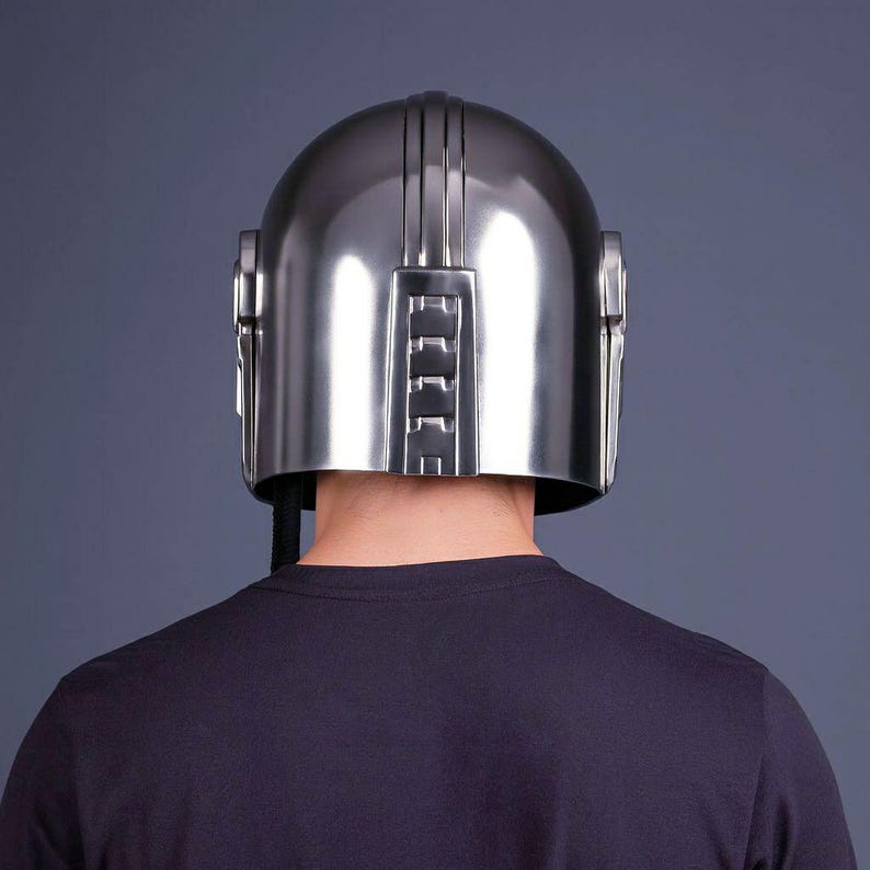 Op de afbeelding: Een zilverkleurige helm met verticale lijnen en rechthoekige details. De helm wordt gedragen door een persoon die een zwart shirt draagt. De helm heeft een metallic glans en een futuristisch ontwerp. Het ontwerp van de helm doet denken aan een sciencefictionpersonage.