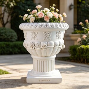 May include: A white, ornate planter filled with pink and white roses. The planter has a detailed design with floral and leaf patterns, and sits on a square base. The text "JINHUOBA" is printed on the base.