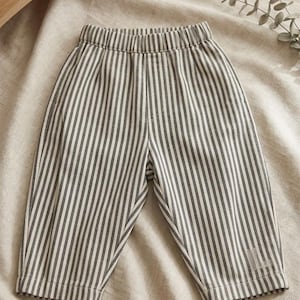 May include: Gray and white striped baby pants. The pants have an elastic waistband and are made of a soft fabric. The pants are displayed on a neutral-colored linen surface.