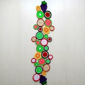 May include: A vertical wall hanging composed of numerous circular crochet pieces resembling various fruit slices, including oranges, limes, watermelons, and kiwis, with purple crocheted grape clusters at the top and bottom ends.