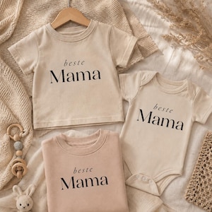 Op de afbeelding: Drie beige baby outfits met de tekst "beste Mama" in het zwart. De outfits omvatten een t-shirt, een onesie en een sweatshirt. Een houten kleerhanger, een houten speelgoed en een knuffelkonijn staan ook op de foto.