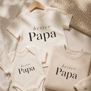 May include: Cream-colored baby bodysuit, t-shirt, and sweatshirt set. Each item features the text "bester Papa" in a black, elegant font. The t-shirt is hanging on a wooden hanger.