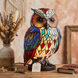 May include: A colorful stained glass owl lamp with a detailed design. The lamp features a variety of colors, including blue, red, yellow, and orange. The lamp is on a dark base and has a tag that says "ARTISAN".