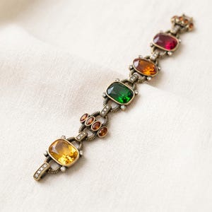 Könnte beinhalten: Ein Armband im antiken Stil mit Bronze-Finish. Das Armband verfügt über eine Reihe von farbigen, rechteckigen Edelsteinen in Gelb, Grün, Orange und Rot. Kleine, klare Kristalle und dekorative Bronzelinks verbinden die Edelsteine. Das Armband ist vor einem weißen Hintergrund platziert.