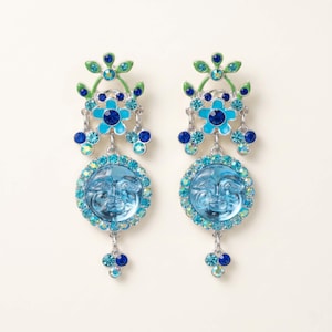 Könnte beinhalten: Ein Paar Ohrhänger mit himmlischem Design. Jeder Ohrring hat einen hellblauen Gesichtsanhänger, der von schillernden Kristallen umgeben ist. Die Ohrringe haben auch ein Blumendesign mit blauen und grünen Akzenten und eine Ansammlung blauer Kristalle am unteren Ende.