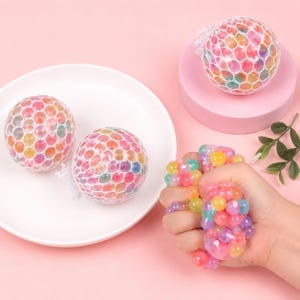 May include: Colorful stress balls filled with small, round, gel-like beads. The balls are encased in a white mesh netting. One ball is being squeezed in a hand, showing the beads. The background is a light pink color.