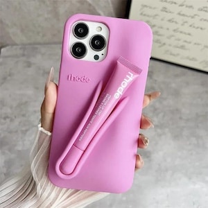 Peut inclure: Une coque de téléphone rose clair avec le nom de la marque "rhode" en relief au dos. Un tube de baume à lèvres rose assorti est fixé à la coque. Le tube de baume à lèvres porte le nom de la marque "rhode".