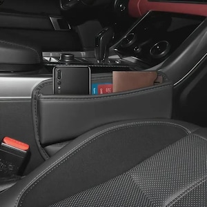 May include: Black car seat organizer with a pocket for a smartphone, a wallet, and cards. The organizer is installed between the car seats and the center console. The interior of the car is black and gray.