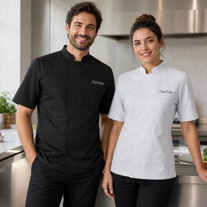 Puede incluir: Una chaqueta de chef negra de manga corta con cuello mao y el texto bordado Chef Andre, junto a una chaqueta de chef blanca de manga corta con cuello mao y el texto bordado Chef Sofia, ambas con un bolsillo en el pecho.