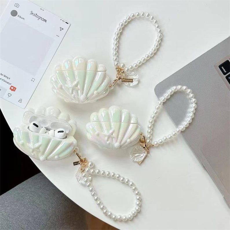 May include: Iridescent white seashell-shaped AirPod cases with pearl wrist straps. One case is open, revealing white AirPods. The cases have a pearlescent finish and a starfish design. The wrist straps are made of small, white pearls.