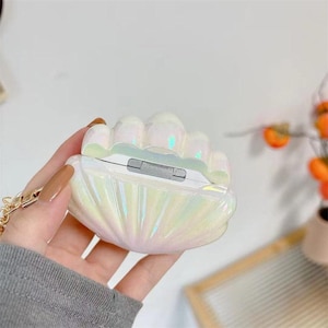 May include: Iridescent seashell-shaped case for wireless earbuds. The pearlescent case has a clam shell design with a gold chain. The case is white with a shimmering, opalescent finish, reflecting various colours. The case is open, revealing the earbuds inside.