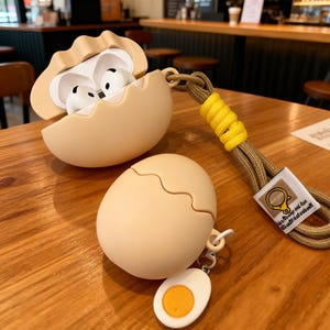 May include: A beige egg-shaped case for wireless earbuds, with a matching eggshell-shaped lid. The case is open, revealing white earbuds. A matching egg-shaped bottom case and a fried egg charm are also present. A brown and yellow lanyard is attached.