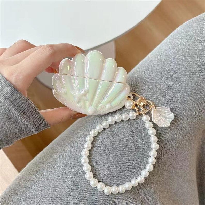 May include: A white, iridescent seashell-shaped case with a pearl bracelet attached. The case has a gold clasp and a small seashell charm. The case is held in a person's hand, resting on grey fabric.