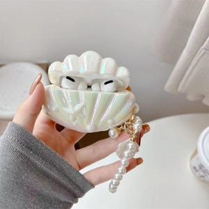 May include: Iridescent white seashell-shaped AirPod case with a pearl charm. The case is open, revealing white AirPods. The case has a pearlescent finish and a pearl and gold charm attached.