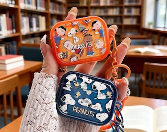 May include: Two AirPods cases with Peanuts characters. One is orange with various characters and the word "PEANUTS". The other is navy blue with Snoopy and the word "PEANUTS". Both cases have a keychain attached for easy carrying.