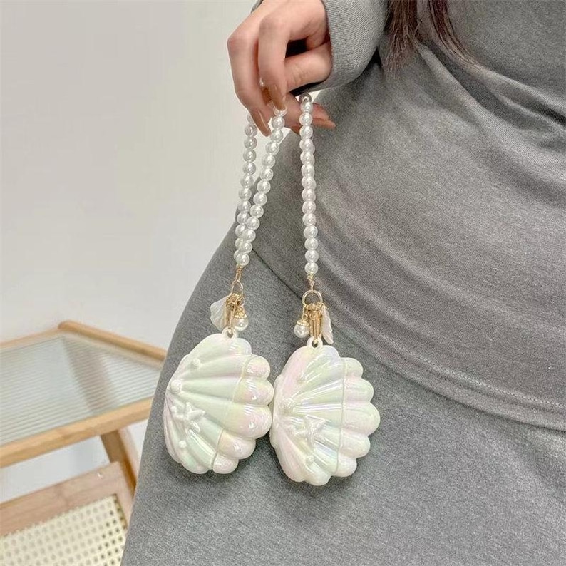 May include: Two iridescent white seashell-shaped accessories with pearl beaded straps. The accessories have gold-coloured accents and are held by a person. The shells have a pearlescent sheen and are detailed with radial lines.