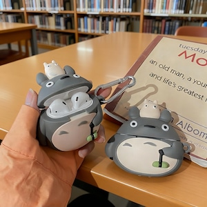 Puede incluir: Funda para AirPod con temática de Totoro gris y blanco con una pequeña figura blanca encima. La funda está abierta, revelando auriculares blancos. Una segunda funda está cerrada.