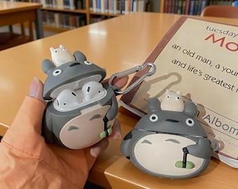 May include: Grey and white Totoro-themed AirPod case with a small white figure on top. The case is open, revealing white earbuds. A second case is closed. The cases are on a wooden table.