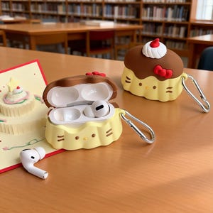 May include: Yellow and brown silicone AirPod cases shaped like cupcakes, with a red bow and white frosting detail. One case is open, revealing white AirPod earbuds. A single AirPod rests on the table. The cases have silver carabiner clips.