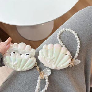 May include: Two iridescent white seashell-shaped AirPod cases. One case is open, revealing white earbuds. Both cases have pearl-beaded straps and gold-coloured accents. The cases are on a person's lap.