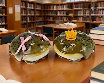 May include: Two ceramic frog figurines with dark green spotted backs and cream-coloured bellies. One frog features two light pink ribbons attached to its head, while the other frog wears a small yellow crown with red dots on its head.