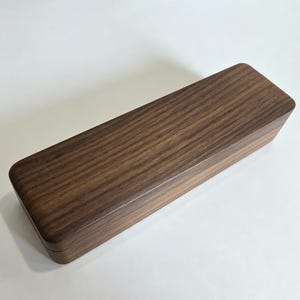 May include: A rectangular wooden box with a dark brown, natural wood grain finish. The box features a smooth, flat top surface and rounded edges, constructed from a single piece of wood with a visible seam where the lid meets the base.