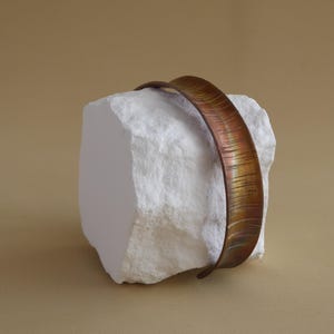 May include: A copper-coloured metal bracelet with a textured surface rests on a white, rectangular stone. The bracelet has a curved design and is displayed against a neutral background.