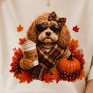 Puede incluir: Una camiseta beige con el gráfico de un perro marrón que lleva gafas de sol y un lazo con estampado de leopardo, una bufanda de cuadros y un collar de perlas. El perro sostiene un vaso con la etiqueta Pumpkin Spice, rodeado de hojas de otoño.