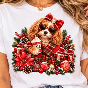 Pode incluir: Uma t-shirt branca com o gráfico de um cão spaniel castanho a usar óculos de sol, um colar de pérolas e um laço vermelho. O cão segura um copo de café no meio de um cenário festivo com ramos de pinheiro, poinsétias vermelhas e presentes.