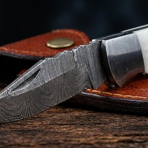 May include: A folding pocket knife with a patterned Damascus steel blade and a white handle secured with metal pins. The knife is positioned partially open, resting against a brown leather sheath with a brass-colored snap button closure.