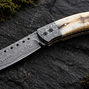 May include: A folding pocket knife featuring a Damascus steel blade with a series of circular holes along the spine. The handle is crafted from smooth, cream-colored bone with dark brown natural grain patterns, secured by three small metal rivets.